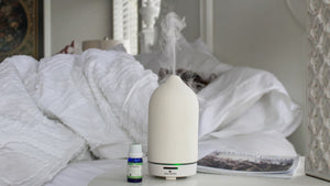 a person in the background snoozes in bed on a rainy day while a stone diffuser in the foreground diffuses essential oils 