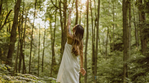 a woman in a white dress reaches one arm up in a sunlit forest 